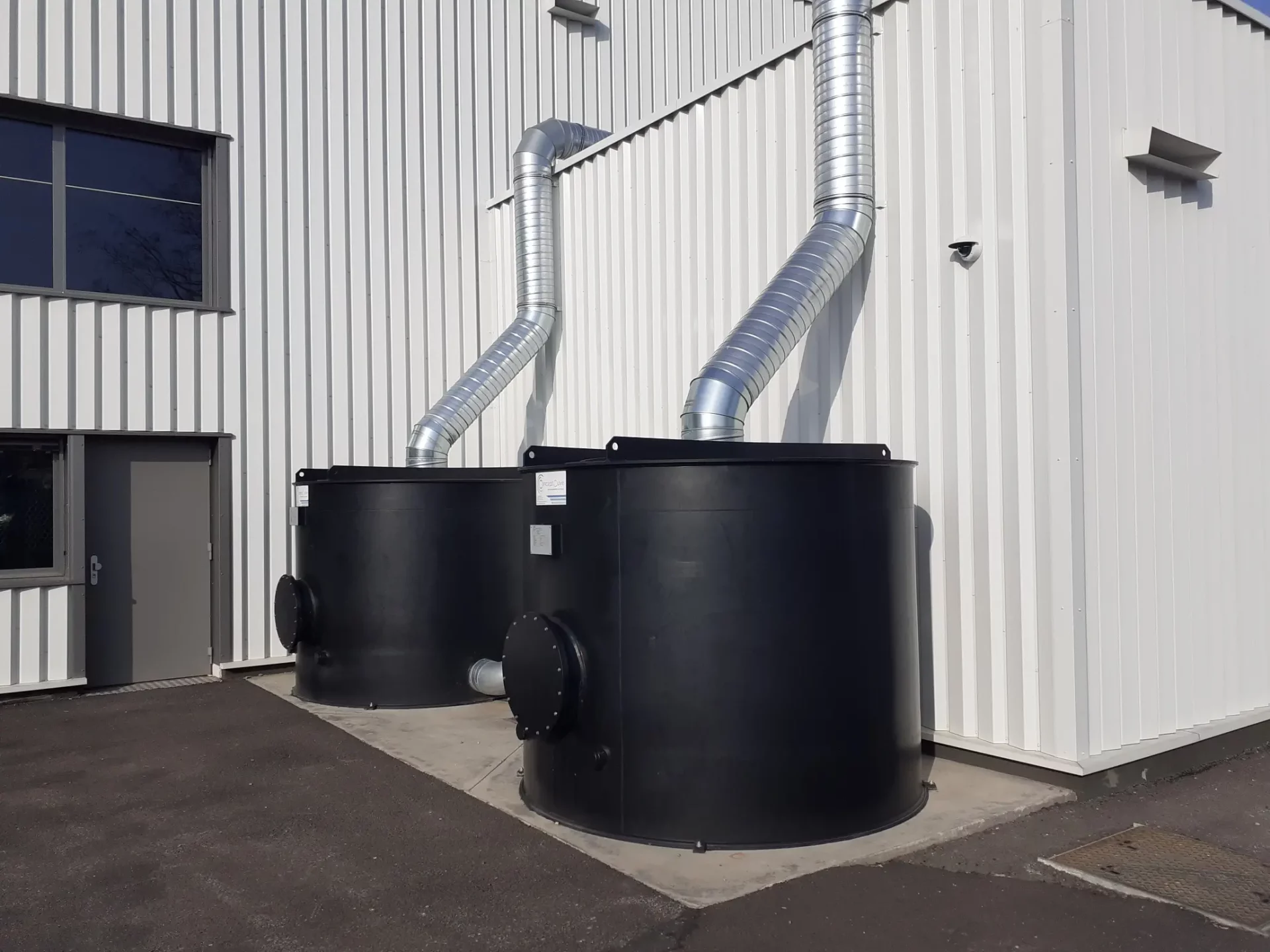 Deux filtres industriels noirs raccordés à des conduits d’extraction d’air, à l’extérieur d’un bâtiment.
